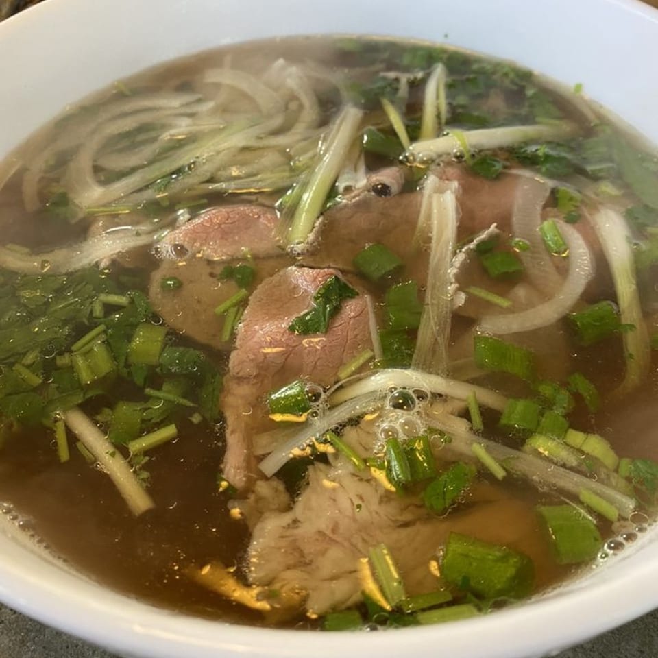 Close-up of I Love Pho's Vietnamese pho with beef, noodles, onions, and green herbs in broth.