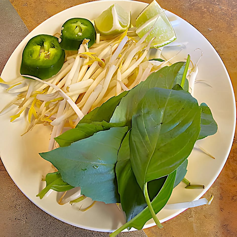 A white plate of fresh pho garnishes: lime wedges, jalapeño slices, bean sprouts, and basil.